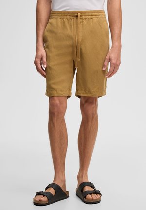 Mężczyzna ubrany w beżowe szorty ze sznurkiem i czarne sandały na sprzączkę, stojący na jasnym, jednolitym tle.