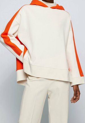 Person wearing off-white hoodie and pants with bright orange stripes along sleeves and hood, standing against plain background.