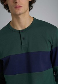 Camicia verde a maniche lunghe con una striscia orizzontale blu navy, colletto rotondo e due bottoni neri sulla patta. Tessuto dalla texture liscia.