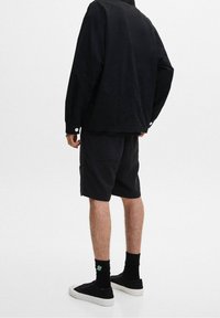 Schwarzes Jacken- und Shorts-Outfit. Die Jacke hat einen entspannten Schnitt mit Knopf-Akzenten, während die Shorts kniehoch mit einer strukturierten Oberfläche sind. Schwarze Sneaker werden getragen.