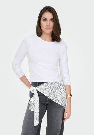 Kvinde med skulderlangt brunt hår iført en hvid langærmet top, mørke jeans og et hvidt blonder tørklæde bundet om taljen.