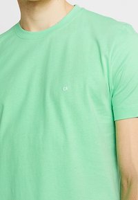 Camiseta de algodón verde con mangas cortas, cuello redondo y pequeño logo blanco "ck" en el lado izquierdo del pecho. Textura suave y ajuste casual.