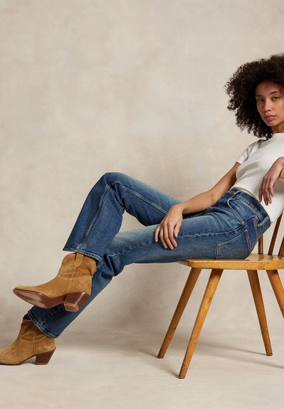 Žena s kudrnatými vlasy, opírající se o dřevěnou židli, oblékající bílé tričko, modré džíny a béžové semišové kotníkové boty na béžovém pozadí.