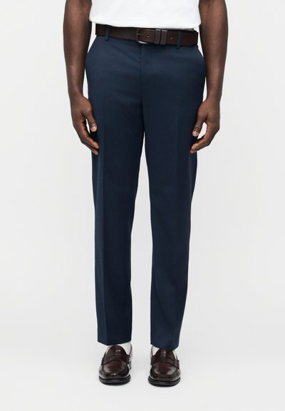 Homme portant un pantalon de costume bleu marine, une chemise blanche rentrée, une ceinture marron foncé, des chaussettes blanches et des mocassins marron foncé, debout devant un fond blanc.