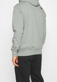 Sudadera verde clara hecha de una tela suave, con un bolsillo estilo canguro, puños y dobladillo acanalados, y un logo en la parte posterior. Combinada con pantalones negros.