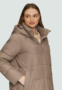 Beige gepolsterte Jacke mit Kapuze, versehen mit horizontalen Nähten, Reißverschluss und seitlichen Taschen. Weiche, glatte Textur mit verstellbaren Kordeln.