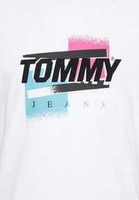 Camiseta blanca con un audaz logo negro "TOMMY", flanqueado por un acento rectangular rosa y azul, con una textura desgastada. "JEANS" en azul claro debajo.