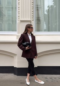 Blazer oversize bordeaux sopra una camicia bianca, leggings neri e ballerine bianche; tiene una pochette nera; in piedi accanto a un muro chiaro.
