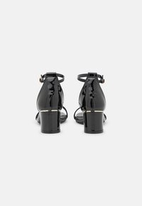 Svart lackskinn klacksandaler med tjock klack och ankelremmar. Har en guldaccent vid basen av klacken. Slät, glansig yta.