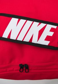 Rode canvas tas met een groot wit "NIKE" logo, zwart trim en dubbele zwarte ritsen aan de onderkant voor sluiting en toegang.