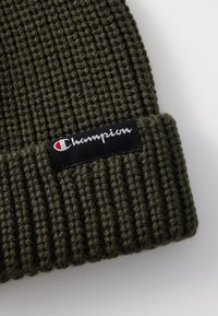 Beanie verde lavorato a maglia con texture a coste. Presenta un'etichetta nera con testo bianco e un accentuazione rossa del logo frontalmente.