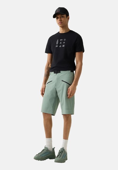 Mladý muž v černém tričku s grafikou, světle zelených šortkách s diagonálními zipovými kapsami, zelených teniskách, bílých ponožkách a černé kšiltovce.