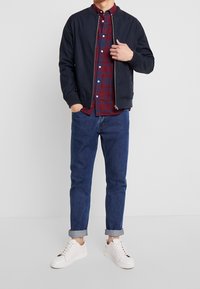 Marine bomberjack over een rood en blauw geruit shirt, gecombineerd met opgerolde blauwe jeans en witte sneakers. Eenvoudig ontwerp, effen kleur en casual pasvorm.