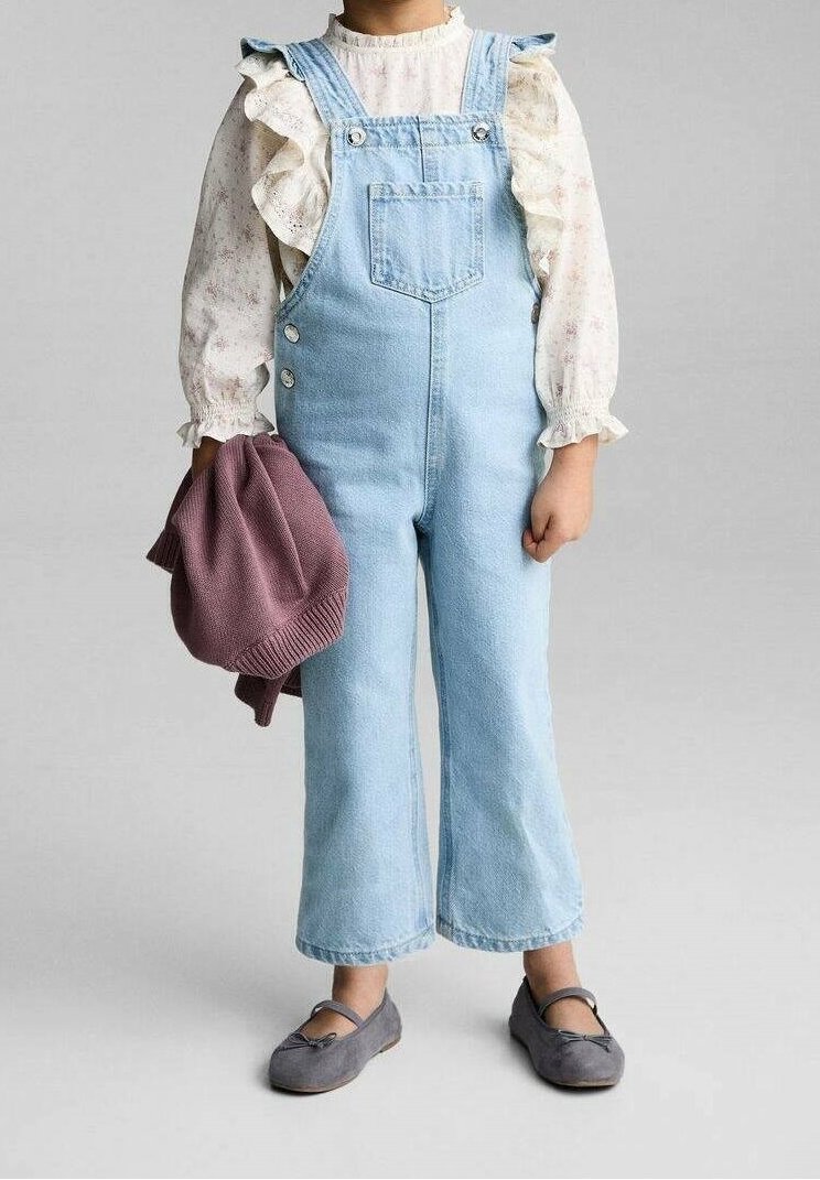 Enfant portant une salopette en denim bleu clair sur un chemisier blanc à manches longues volanté, tenant un pull violet plié, chaussures plates grises aux pieds.