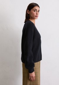 Schwarzer Strickpullover mit gerippter Textur und tiefem V-Ausschnitt. Lange, puffärmel ergänzen die lockere Passform, kombiniert mit einem karierten Rock.