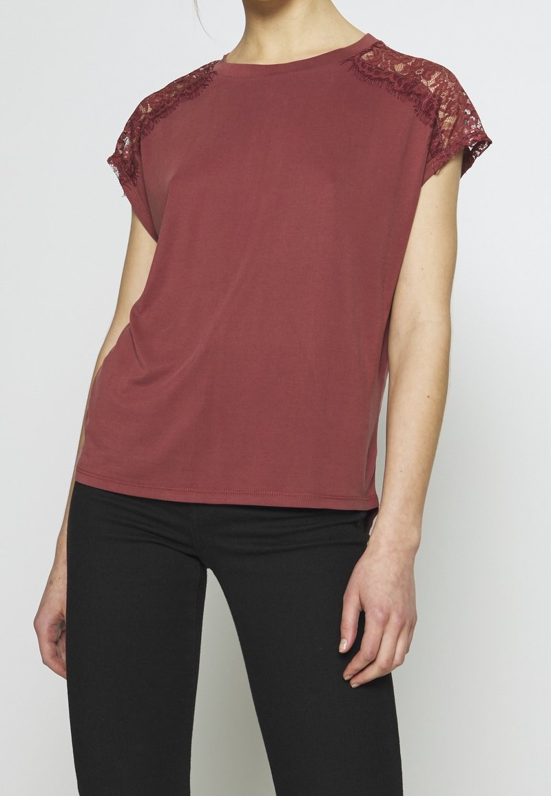 Vínové tričko s krajkovými detaily na ramenou, krátkými rukávy a volným střihem. Hladká látka s kulatým výstřihem. Nošeno s černými kalhotami.