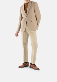 Completo beige con due bottoni, rever a lancia e tasche laterali. Maglione color beige chiaro sotto. Pantaloni beige chiaro e scarpe in pelle marrone con dettagli blu.