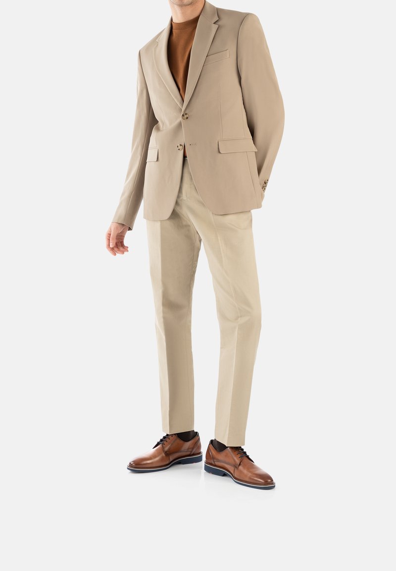 Completo beige con due bottoni, rever a lancia e tasche laterali. Maglione color beige chiaro sotto. Pantaloni beige chiaro e scarpe in pelle marrone con dettagli blu.