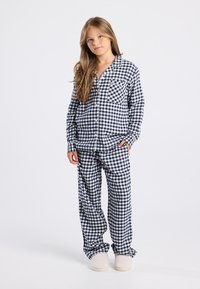 Jong meisje staat in navy en wit geblokte pyjama en crèmekleurige slippers tegen een effen grijze achtergrond.