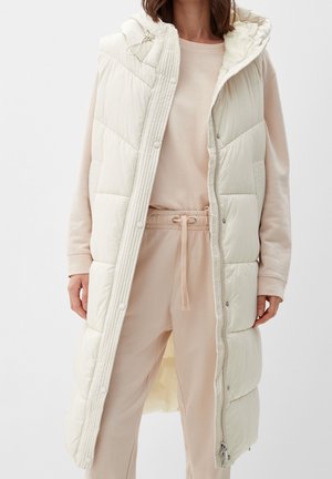 Žena nosící dlouhou béžovou prošívanou vestu s kapucí přes béžovou mikinu a kalhoty se stahovací šňůrkou, stojící před jednobarevným pozadím.