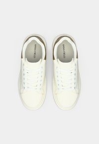 Balts šņorējamos sporta apavos ar zelta akcentiem uz papēžiem, skatīts no augšas, uz vienkārša gaiša fona.