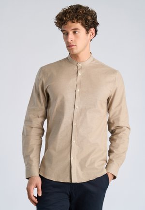 Jonge man met krullend haar, gekleed in een beige overhemd met mandarin kraag en donkere broek, kijkt opzij met één hand in zijn zak.