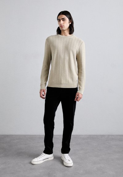 Pull en maille beige avec un motif en chevron texturé, associé à un pantalon noir et des tennis blanches, porté par un modèle masculin debout.