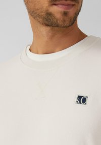 Sudadera color crema con cuello redondo, puños acanalados y un pequeño logo en azul marino en el lado izquierdo del pecho. Textura suave, diseño casual.