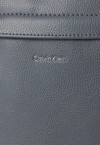 Γκρι υφή δερμάτινης επιφάνειας με ανάγλυφο λογότυπο "Calvin Klein" σε ασημί χρώμα. Το υλικό έχει υφή με κοκκώδες φινίρισμα και ραμμένες λεπτομέρειες.