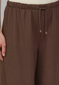 Pantalons larges marron texturés avec taille à cordon et accents métalliques. Le tissu semble doux et léger, créant une coupe décontractée.