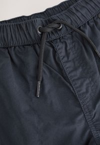 Primo piano di pantaloncini con fascia elastica nera in vita e cordoncino nero legato a nodo al centro davanti.