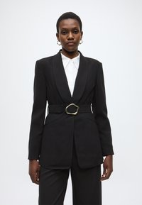 Μαύρο σακάκι με γιακά notch, μέση με ζώνη και χρυσή αγκράφα, και δύο μπροστινές τσέπες. Φορεμένο πάνω από ένα λευκό πουκάμισο με κουμπιά.