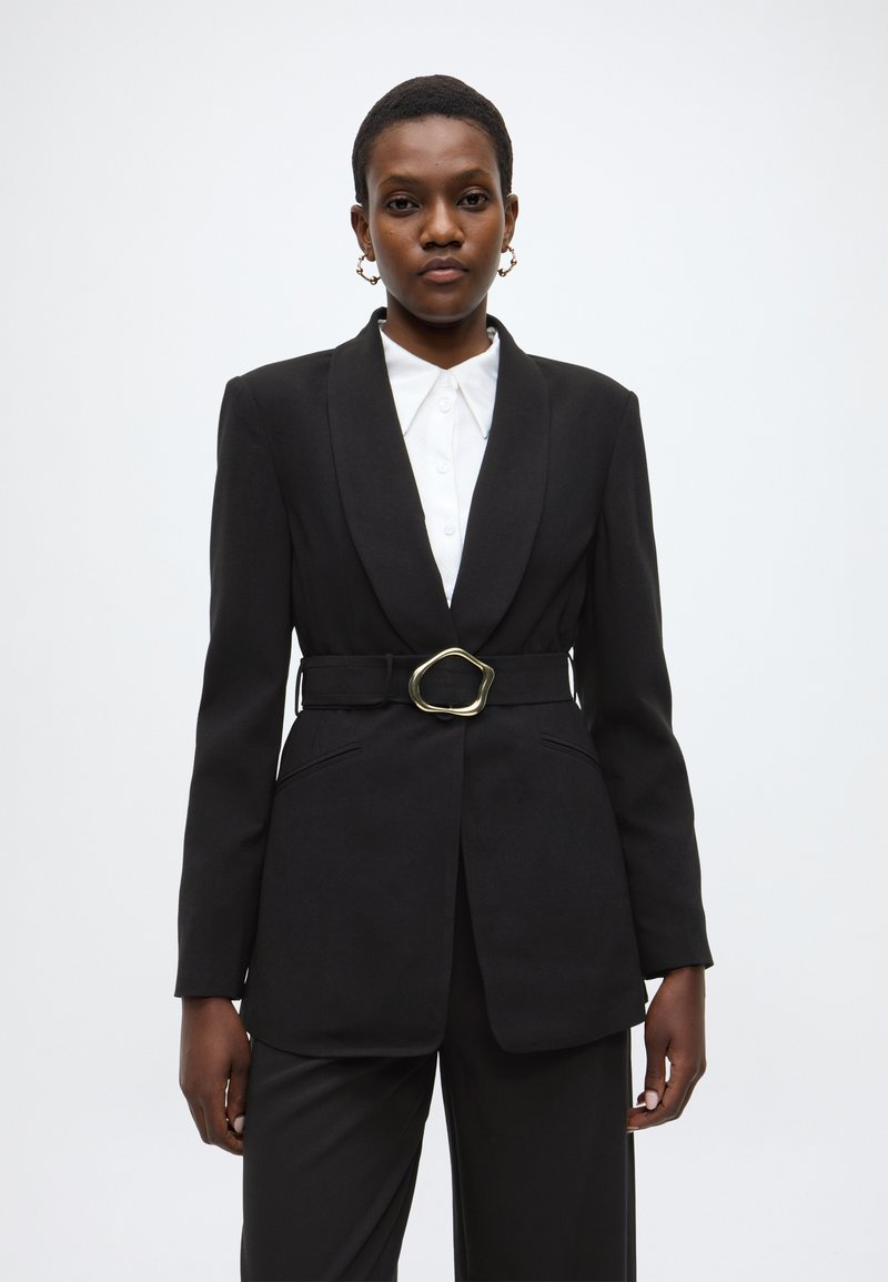 Μαύρο σακάκι με γιακά notch, μέση με ζώνη και χρυσή αγκράφα, και δύο μπροστινές τσέπες. Φορεμένο πάνω από ένα λευκό πουκάμισο με κουμπιά.