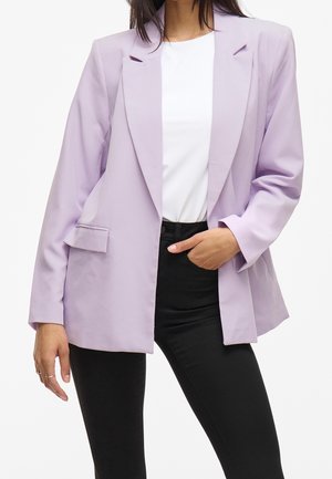 Frau trägt einen hellvioletten Blazer über einem weißen Hemd mit schwarzen Hosen, eine Hand in der Tasche, steht vor einem einfarbigen Hintergrund.
