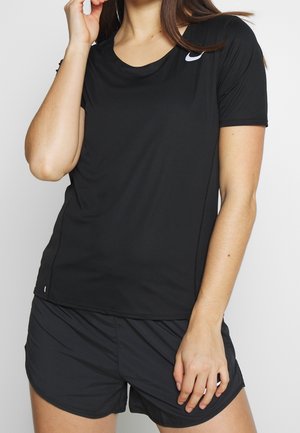 Femme portant un t-shirt noir Nike à manches courtes et un short de sport noir, une main près du visage et l'autre reposant sur sa cuisse.