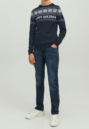 Personne portant un pull de fête bleu marine avec des motifs de flocons de neige blancs et le texte « Joyeuses Fêtes », un jean bleu et des baskets blanches.