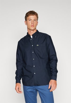 Navy blue button-up shirt with a collar, long sleeves, and green logo on the chest pocket; made of smooth fabric with a solid texture.
