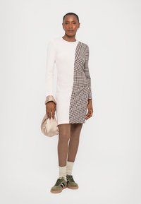 Rochie în două tonuri, cu țesătură texturată de culoare roz pal pe o parte și țesătură pătrată maro pe cealaltă. Însoțită de o poșetă bej și teniși verzi olive.