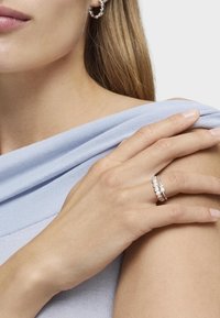 Srebrny pierścionek z wieloma drobnymi białymi kamieniami szlachetnymi, gładka faktura, noszony na dłoni spoczywającej na jasnoniebieskiej tkaninie.