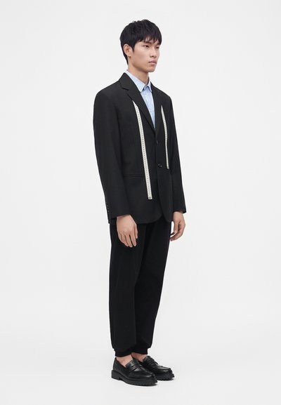 Jeune homme portant une veste de costume noire avec un mètre ruban autour du cou, pantalon noir et chaussures noires, debout sur un fond blanc.