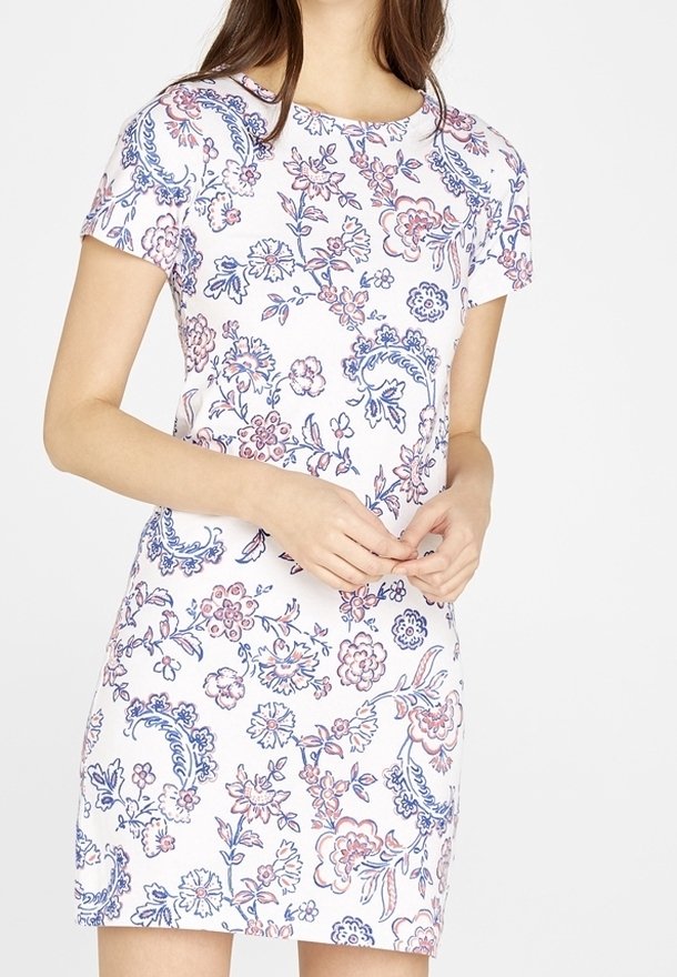 Femme portant une robe blanche à manches courtes avec des motifs floraux roses et bleus, mains jointes à la taille, debout devant un fond blanc uni.