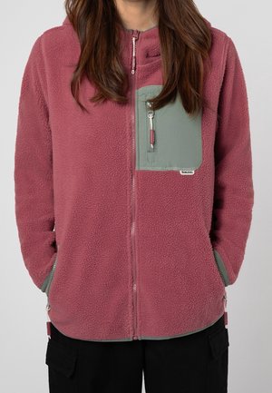 Veste en polaire rose poudré avec une poche zippée vert clair, un col montant et des fermetures éclair argentées. Comprend des poches latérales et un tissu texturé.