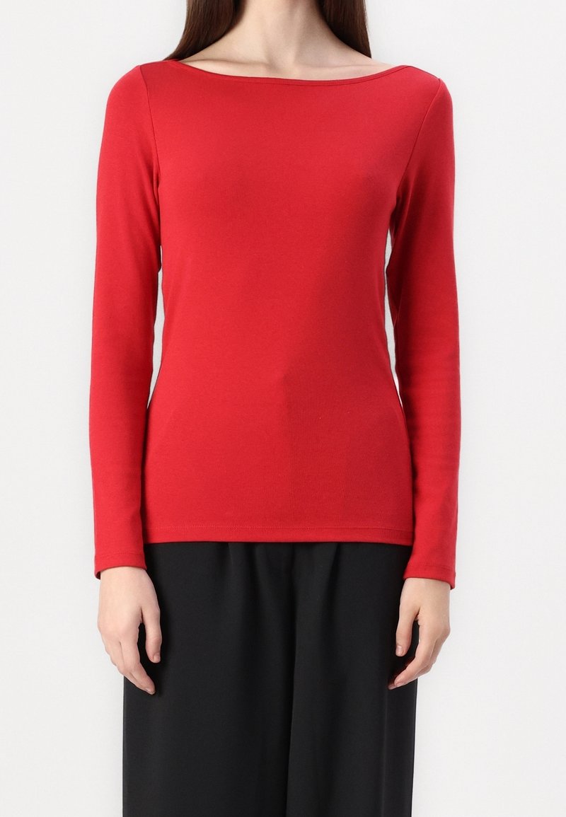 Femme portant un haut rouge uni à manches longues avec un large col bateau et un pantalon noir ample, debout devant un fond blanc.