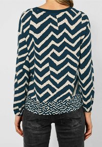 Blusa a maniche lunghe con base color teal e motivi a zigzag bianchi, con un orlo decorato. Realizzata in un tessuto morbido e leggero.
