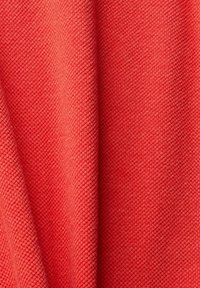 Kankainen kangas kirkkaassa korallivärissä, jossa on kiinteä kudontamalli hienovaraisella kiillolla, laskeutuen sujuvasti pehmeältä ja paksulta vaikuttavaltä.