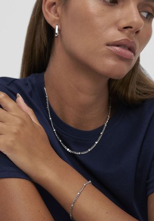Femme portant des boucles d'oreilles créoles en argent, un collier en chaîne argenté, un bracelet assorti, et une chemise bleu marine, avec le bras croisé sur sa poitrine.