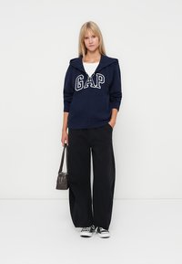 Marinblå huvtröja med "GAP"-logotyp, framzipper, avslappnad passform, kombinerad med vida svarta byxor och en vit t-shirt, kompletterad med sneakers.