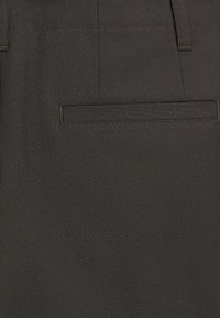 Detail kapsy ze zadní části tmavě šedé látky na šitých kalhotách s viditelným prošitím a poutky na opasek.