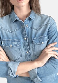 Chemise en denim bleu clair avec deux poches poitrine, boutons-pression et coutures contrastées. Elle possède un col pointu et des manches retroussées.