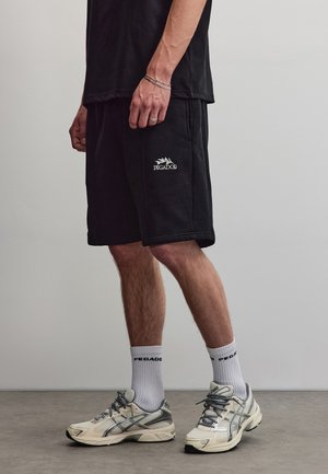 Hombre con pantalones cortos Pegador negros, calcetines Pegador blancos y zapatillas deportivas blancas y grises, de pie sobre un suelo gris frente a una pared lisa.